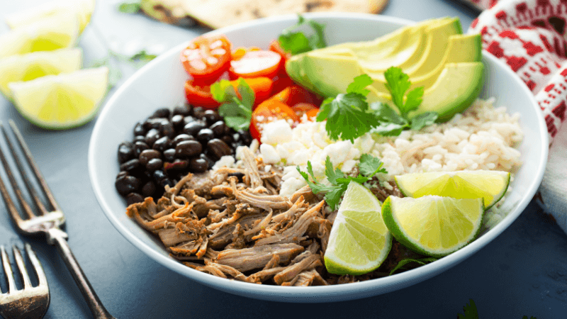 a white burrito bowl filled with black beans, rice, sliced avocados, shredded chicken, halved cherry tomatoes & lime wedges, garnished with cilantro