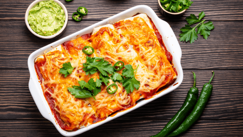 a white tray with cheesy baked enchilada topped with cilantro served with guacamole & green chillies in rustic wooden table
