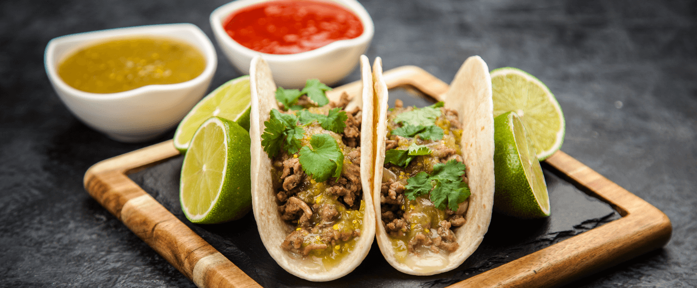 beef tacos served on a wooden board with tomato salsa, salsa verde & halved limes