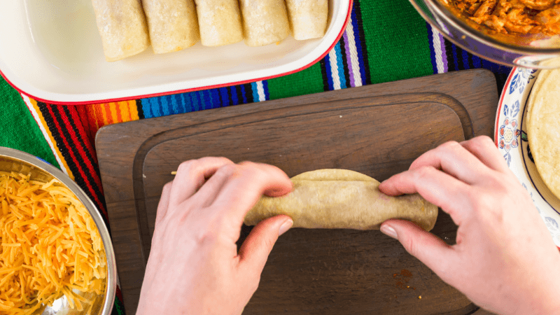 hands rolling enchiladas on wooden board surrounded by cheese & enchiladas in a tray