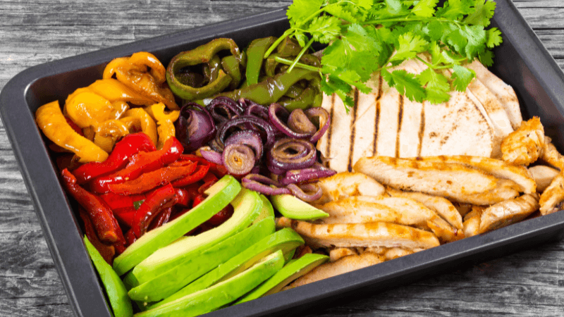 a tray with grilled chicken strips, tortillas, bell peppers, onions, avocado slices & cilantro on a wooden surface