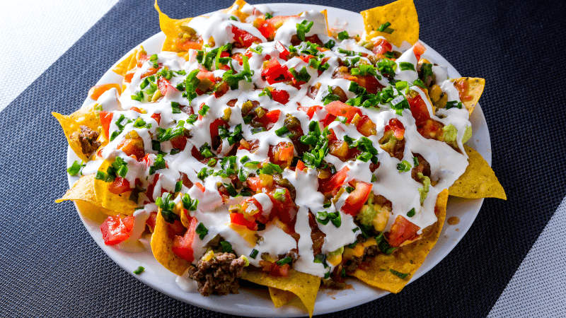 cheesy baked nachos topped with sour cream, diced tomatoes, coriander & other garnishes, served in a white plate on a black background