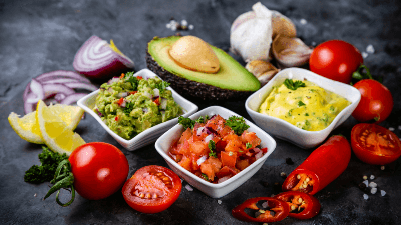 Mexican sauces & toppings such as guacamole, salsa, cheese sauce, sliced onions & tomatoes, chilies, avocado placed on rustic black background