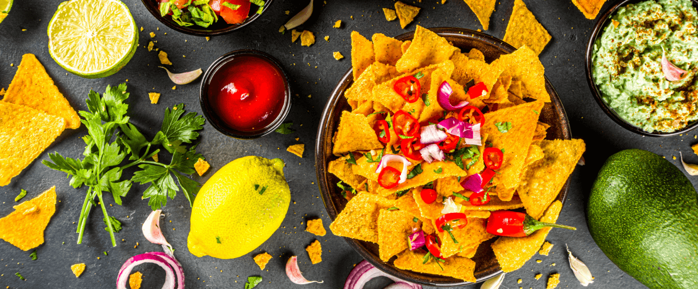 a plate of nachos topped with diced onions, chillies & tomatoes, surrounded by dips, lemon & scattered chips