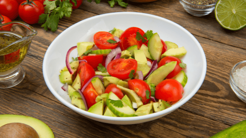 a white bowl filled with a fresh tomato avocado salad, garnished with sliced red onions & herbs, placed on rustic wooden table with ingredients in the background