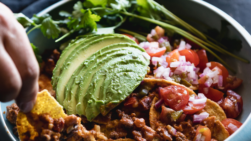 lamb nachos garnished with sliced avocado, diced onions, tomatoes & coriander in a blue pot