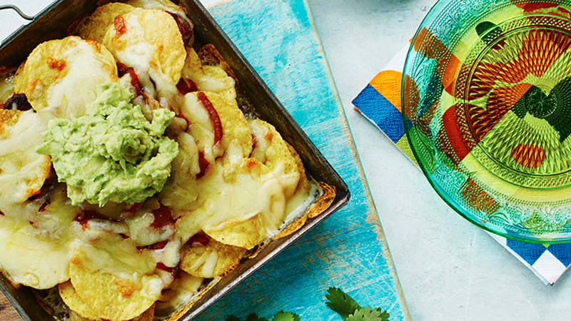 Chorizo Nachos topped with mashed Avocado served in a black square bowl on a rustic blue board