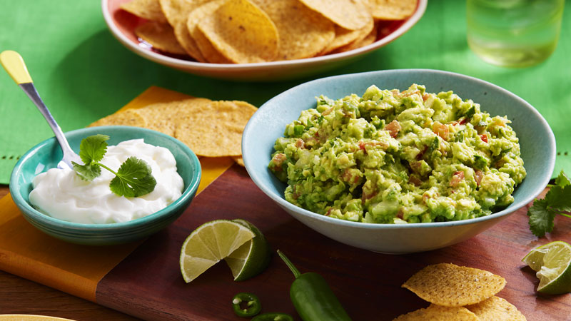 traditional guacamole served in a light blue bowl placed on wooden board, surrounded by sour cream, lime wedges & nachips